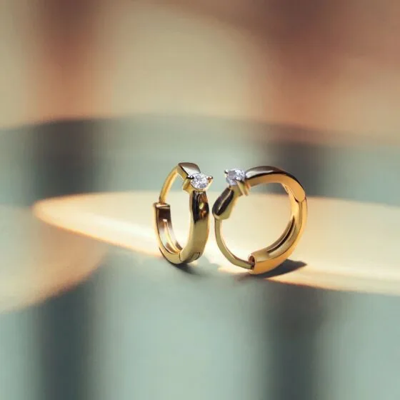 Pair of gold huggie hoop earrings, each set with a single sparkling diamond, standing on a reflective surface.