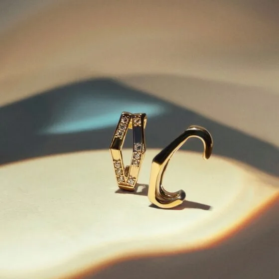 Two modern gold ear cuffs, one plain high-polish and one paved with small crystals, on a beige surface under a warm light.