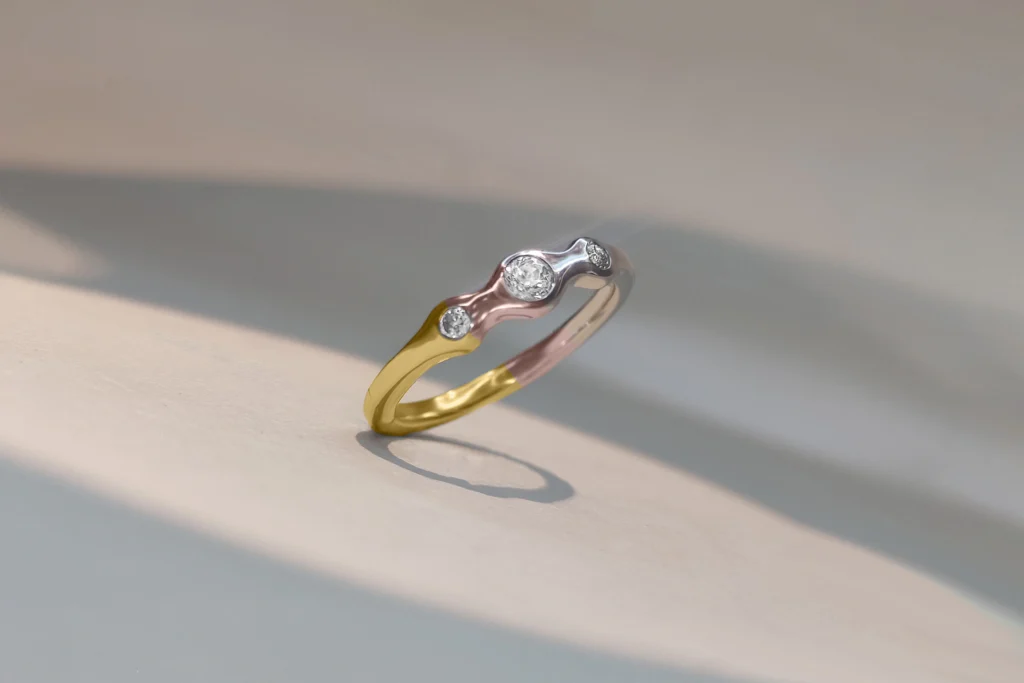 Three-stone tricolor ring featuring yellow, rose, and white bands set with sparkling round diamonds on a soft gray surface.