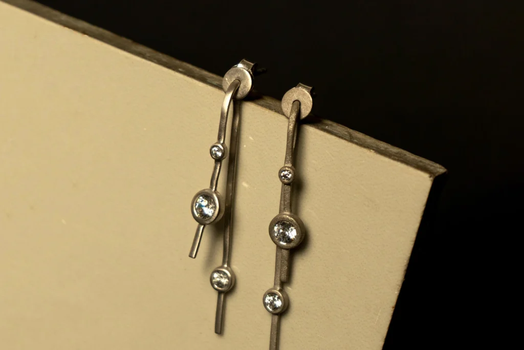 Pair of long matte silver drop earrings with bezel-set round crystals hanging from a textured cream stand.